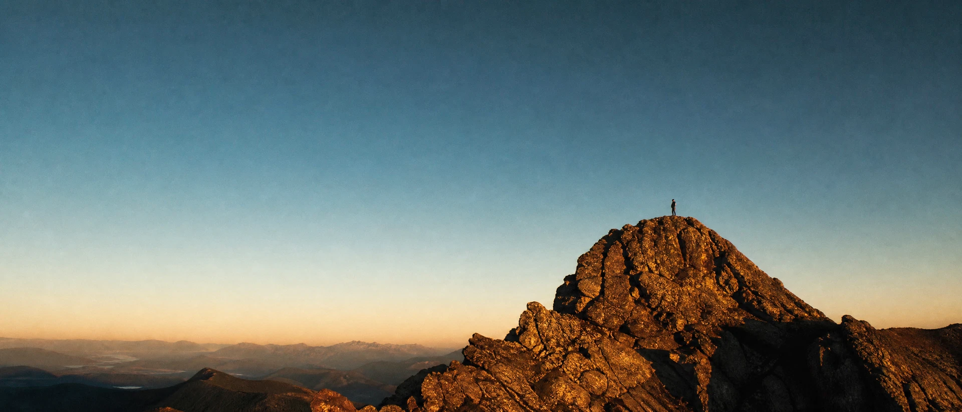 A lone figure standing at the summit of a sunlit rocky outcrop, silhouetted against a vast deep-teal dusk sky