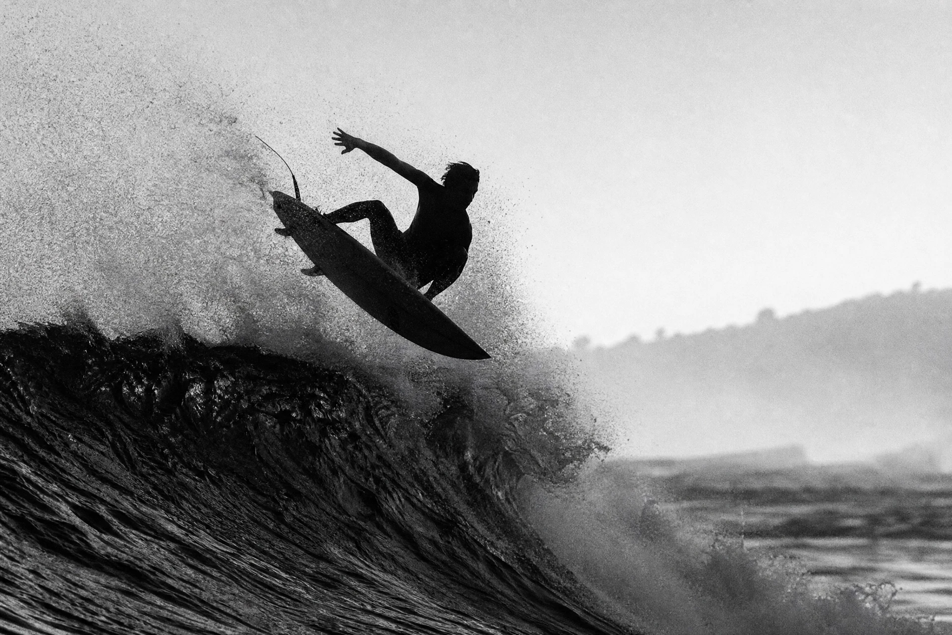 High-contrast black and white photograph of a lone surfer driving off the lip of a large breaking wave